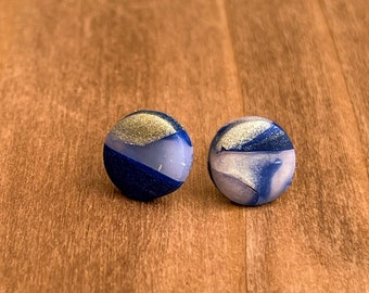 May include: A pair of blue and gold stud earrings with a marbled design. The earrings are on a wooden surface.