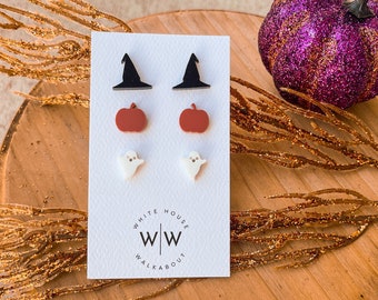 May include: A set of three pairs of Halloween-themed stud earrings. The earrings feature a black witch hat, a red pumpkin, and a white ghost. The earrings are displayed on a white card with the text "White House Walkabout WW".
