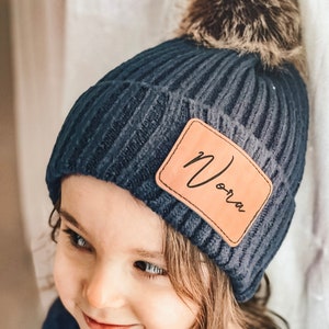 Op de afbeelding: Een donkerblauwe gebreide beanie met een nep bont pompon en een bruin leren patch met de tekst "Nora".