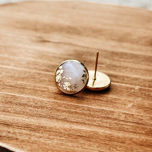 May include: Round stud earrings with a gold-colored border. The earrings have a light-colored center with gold leaf accents. One earring is in focus, while the other is slightly out of focus. The earrings are on a wooden surface.