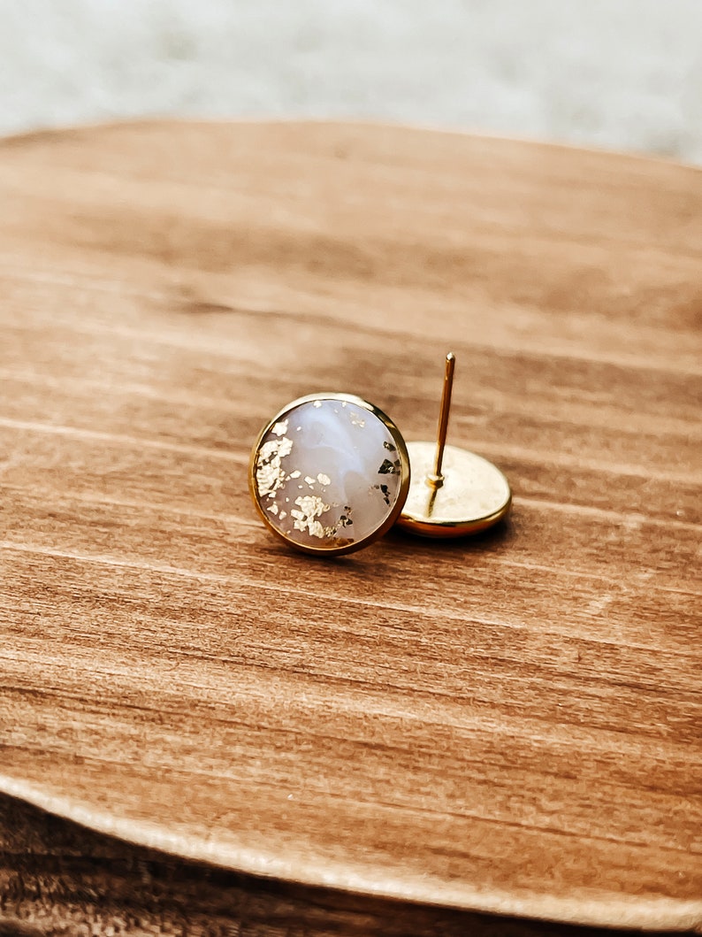 May include: Round stud earrings with a gold-colored border. The earrings have a light-colored center with gold leaf accents. One earring is in focus, while the other is slightly out of focus. The earrings are on a wooden surface.