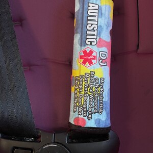 K&ouml;nnte beinhalten: Ein bunter Sicherheitsgurtbezug mit einem Puzzle-Design und dem aufgedruckten Text "AUTISTIC DJ". Der Text unter dem Design lautet "Kann Hilfe verweigern oder nicht reagieren. Risiko des Umherwanderns. Kann Gefahren nicht erkennen. Mag keine lauten Ger&auml;usche."