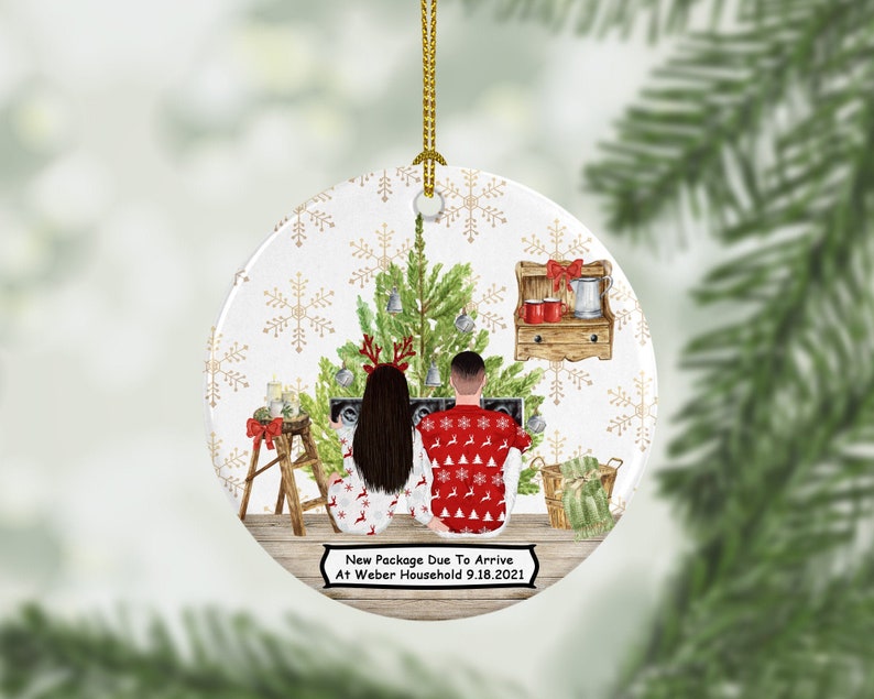 Pu&ograve; includere: Un addobbo natalizio rotondo con sfondo bianco e fiocchi di neve dorati. Una coppia con maglioni natalizi si trova davanti a un albero di Natale decorato. Il testo sull'addobbo dice "New Package Due To Arrive At Weber Household 9.18.2021".