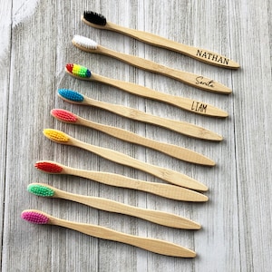 Puede incluir: Nueve cepillos de dientes de bambú personalizados con cerdas de colores. Los cepillos de dientes están dispuestos en una fila sobre una superficie de madera. Los nombres "Nathan", "Santa" y "Liam" están grabados en los cepillos de dientes.