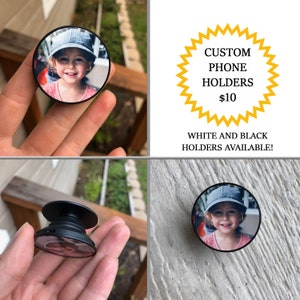 May include: A black phone holder with a round, expandable base. The holder has a custom photo of a young girl wearing a blue hat. The text "CUSTOM PHONE HOLDERS $10 WHITE AND BLACK HOLDERS AVAILABLE!" is visible in the image.