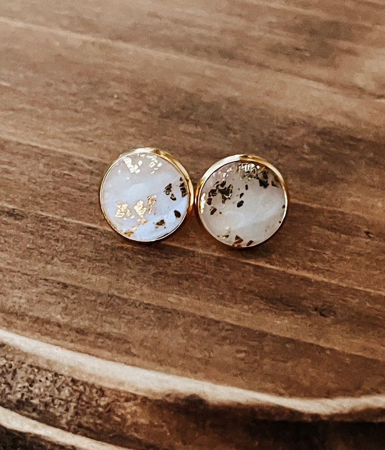 May include: A pair of round stud earrings with a gold-colored border. The earrings have a white background with gold leaf accents. The earrings are displayed on a wooden surface.