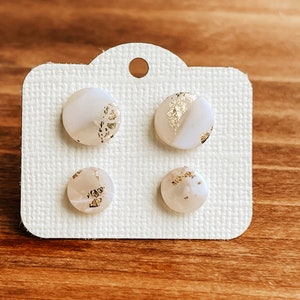 May include: Four white round stud earrings with gold flecks. The earrings are displayed on a white card with a hole at the top.