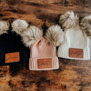 Könnte beinhalten: Drei Strickmützen mit Kunstfellbommeln. Jede Mütze hat einen personalisierten Leder-Patch mit den Namen "Gracelyn", "Daisy" und "Jamie".