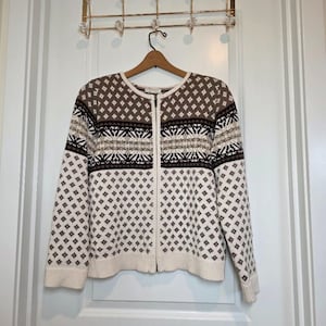 May include: A long-sleeve zip-up cardigan sweater featuring a cream base color with a repeating brown and dark brown geometric diamond pattern. The garment includes a horizontal band across the chest with a detailed snowflake-inspired design.
