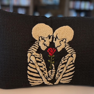 Peut inclure: Un coussin rectangulaire noir orné d'une broderie représentant deux squelettes se faisant face et tenant une rose rouge unique avec des feuilles vertes. Le coussin est agrémenté de franges de couleur crème sur les deux bords latéraux.