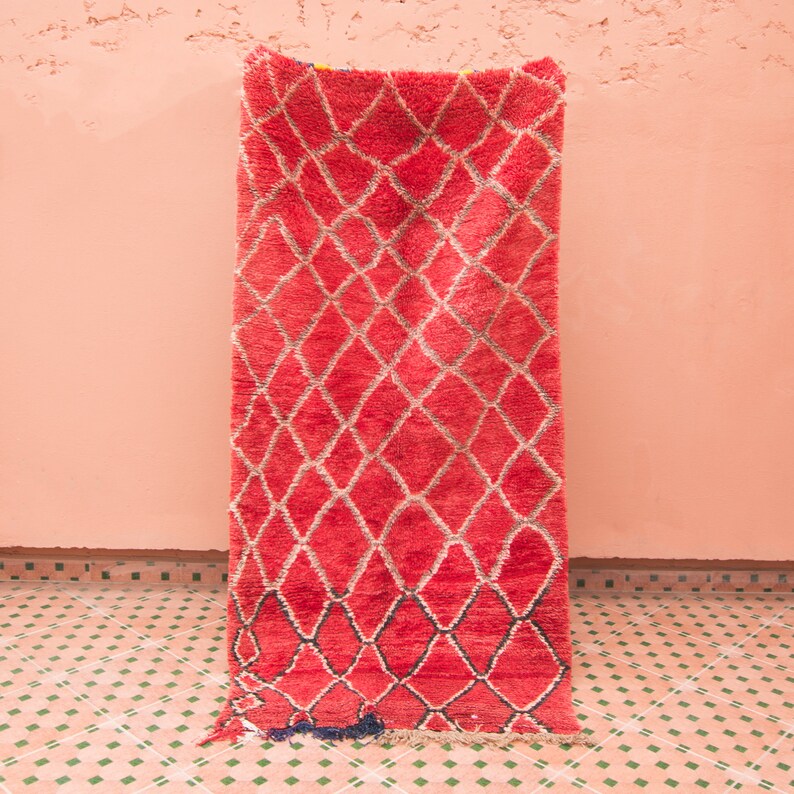 May include: A red Moroccan rug with a diamond pattern. The rug is hanging on a pink wall.