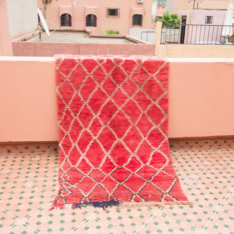 May include: A red and white Moroccan rug with a diamond pattern hangs on a pink wall. The rug has a fringe border and is made of wool.