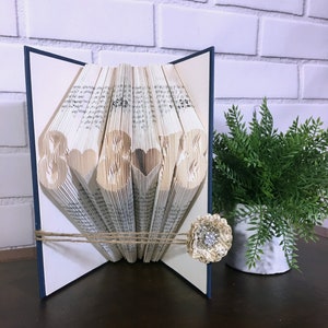 May include: A handmade book art piece with the numbers "8 & 8 & 8" cut out, with heart-shaped cutouts between the numbers. The book is tied with twine and adorned with a decorative flower. The book is displayed on a dark surface.
