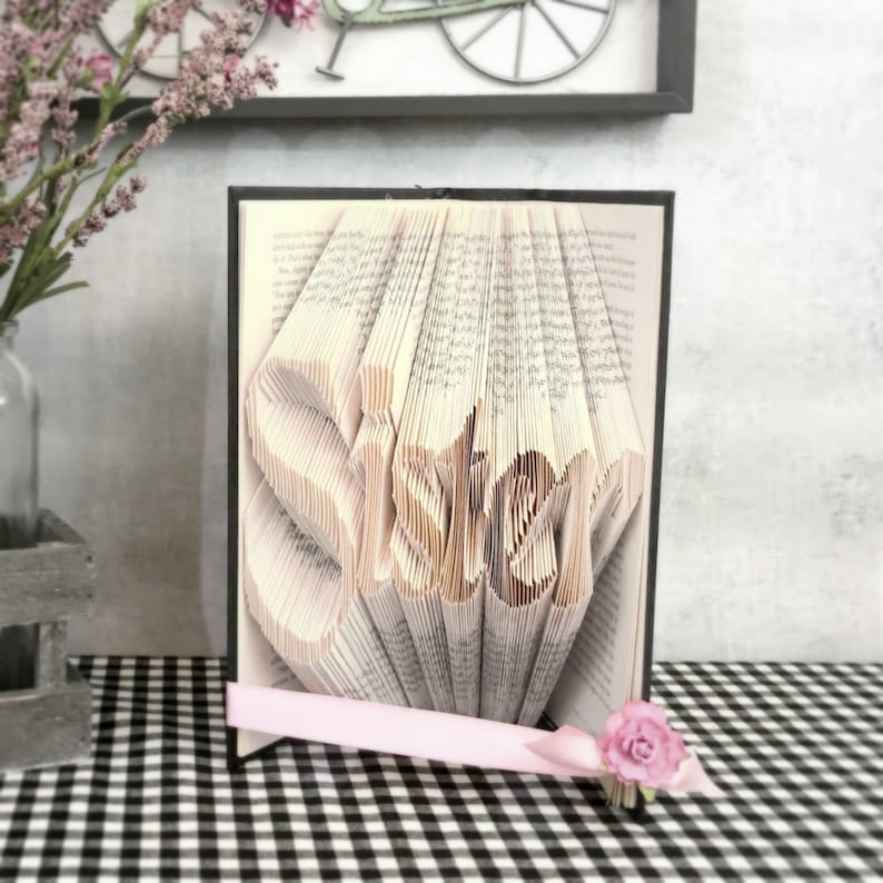 May include: A handmade book art piece with the word "Sister" carved into the pages. The book is open, revealing the sculpted word, and is accented with a pink ribbon and a small pink rose. The background is a black and white checkered pattern.