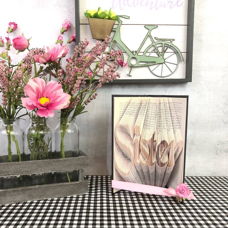 May include: A decorative display featuring a book art piece with the word "Sister" carved into the pages. Pink flowers and a pink ribbon accent the book. A wooden crate holds glass bottles with pink flowers. A sign with a bicycle is in the background.