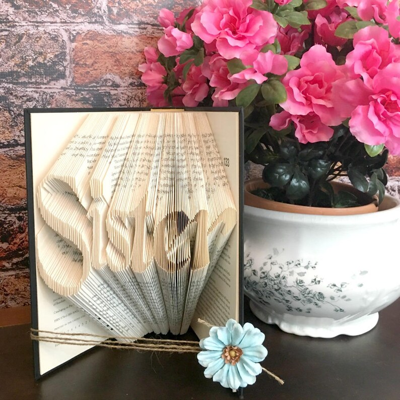 May include: A handmade book art piece with the word "Sister" folded into the pages. The book is black-framed and tied with twine, with a light blue flower and pink azalea plant in the background.