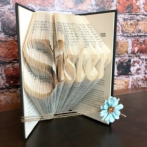 May include: A book sculpture with the word "SISTER" cut into the pages. The pages are folded to create the letters, and the book is tied with twine. A light blue flower is attached to the book.
