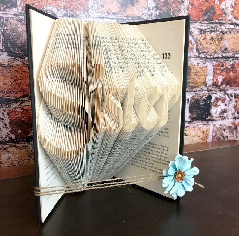 May include: A book sculpture with the word "SISTER" cut into the pages. The pages are folded to create the letters, and the book is tied with twine. A light blue flower is attached to the book.