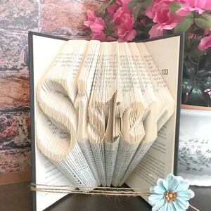 May include: A book sculpture with the word "SISTER" folded into the pages. The book is black with a twine accent and a light blue flower. Pink flowers are in the background.