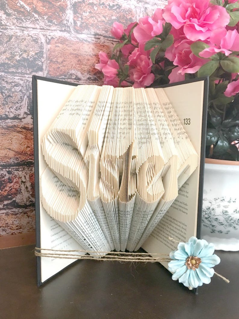 May include: A book sculpture with the word "SISTER" folded into the pages. The book is black with a twine accent and a light blue flower. Pink flowers are in the background.
