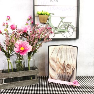 May include: A framed wooden sign with the text "life is an adventure" above a green bicycle with a basket. A wooden crate holds three glass bottles with pink flowers. A book art piece spells "sister".