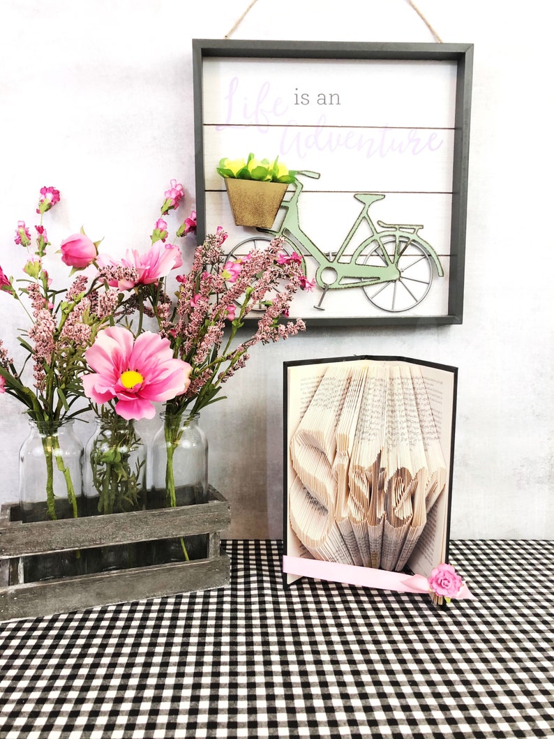 May include: A framed wooden sign with the text "life is an adventure" above a green bicycle with a basket. A wooden crate holds three glass bottles with pink flowers. A book art piece spells "sister".