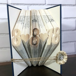 May include: A handmade book art piece with the numbers "8 & 8" folded into the pages. The book is cream-colored with a navy blue spine and is tied with twine. A decorative paper flower with a rhinestone center is attached.