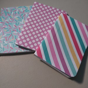 May include: Three small notebooks with different patterned covers. The first notebook has a light blue cover with white and pink sprinkles. The second notebook has a pink cover with white polka dots. The third notebook has a pink, purple, and teal striped cover.