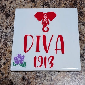 May include: White ceramic tile with a red elephant silhouette, the letters "ΔΣΘ" inside the elephant, the word "DIVA" in red, and the year "1913" in red. A purple flower with green leaves is in the bottom left corner.