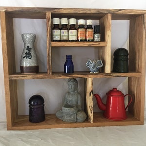 May include: A wooden shelf unit with multiple compartments, featuring a variety of decorative items, including a small gray Buddha statue, a blue glass bottle, a ceramic vase with Japanese characters, a small elephant figurine, a purple glass bottle stopper, a black bottle stopper, and a red teapot.
