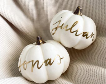 May include: Two white decorative pumpkins with gold lettering. One pumpkin has the name "Jonas" and the other has the name "Julian". The pumpkins have brown stems and are set on a cream-colored textured fabric.