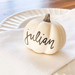 May include: A small, off-white pumpkin-shaped place card holder with the name "julian" written in black script. The pumpkin sits on a white napkin next to a white plate, perfect for fall or Thanksgiving decor.