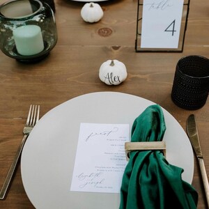 May include: A table setting with a white plate, green napkin, and gold napkin ring. A menu card and a small white pumpkin with the name "Alex" are on the plate. A candle in a glass holder and a table number are also visible.