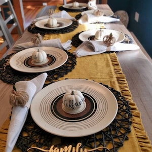 May include: A beautifully set dining table with white plates, black placemats, and a mustard-colored runner. Each place setting includes a small white pumpkin with a name tag. A wooden sign says "family".