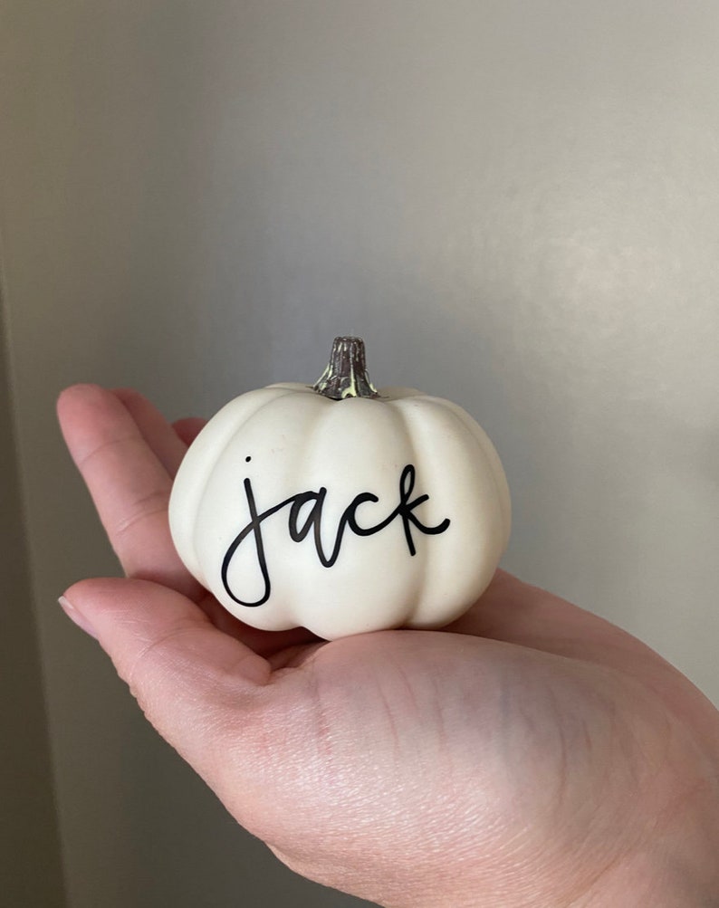 May include: A small, ivory-colored pumpkin with the name "jack" in black script. The pumpkin has a textured surface and a brown stem. A decorative item for fall or Halloween.