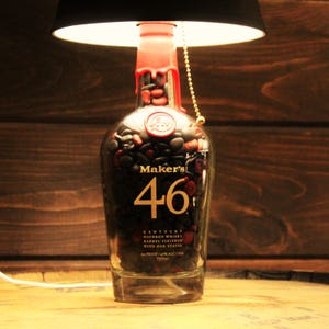 May include: A unique lamp made from a Maker's 46 bourbon whiskey bottle. The bottle is filled with small, dark stones and sealed with red wax. The lamp has a black lampshade and a pull chain. The bottle reads "Maker's 46" and other text.