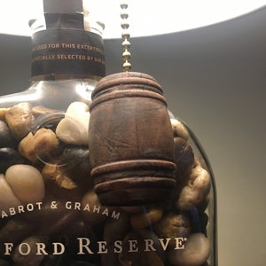 May include: A clear glass bottle filled with small rocks, with a wooden barrel-shaped pull chain hanging from the top. The bottle is labeled "Labrot & Graham Woodford Reserve".