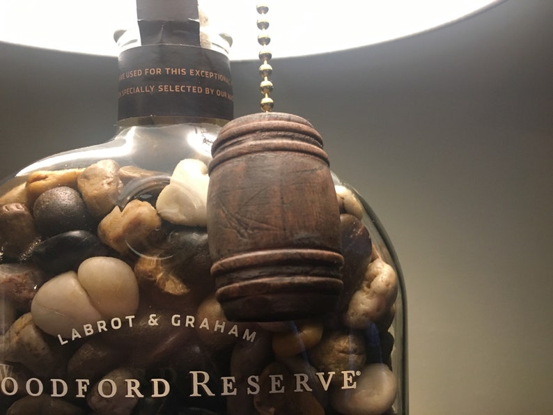 May include: A clear glass bottle filled with small rocks, with a wooden barrel-shaped pull chain hanging from the top. The bottle is labeled "Labrot & Graham Woodford Reserve".