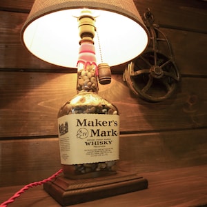 May include: A unique lamp crafted from a Maker's Mark whisky bottle, filled with small stones. The lamp features a brown lampshade, a wooden base, and a red electrical cord. A small wooden barrel is attached to the bottle.