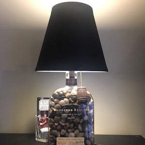 May include: A black lampshade sits atop a clear glass bottle filled with small rocks. The bottle is labeled "Woodford Reserve Distiller's Select Kentucky Straight Bourbon Whiskey".