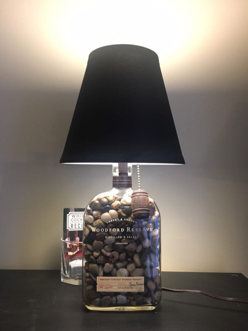 May include: A black lampshade sits atop a clear glass bottle filled with small rocks. The bottle is labeled "Woodford Reserve Distiller's Select Kentucky Straight Bourbon Whiskey".