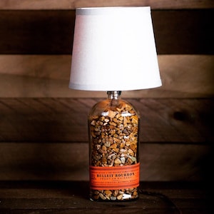 May include: A table lamp made from a clear glass bottle filled with small brown stones. The bottle has an orange label that reads "BULLEIT BOURBON FRONTIER WHISKEY". The lamp has a white lampshade.
