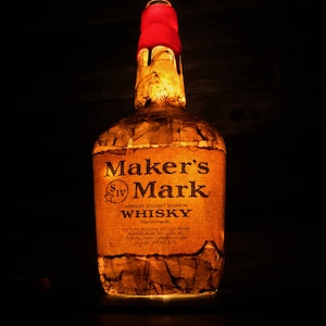 May include: A glass bottle of Maker's Mark Kentucky Straight Bourbon Whisky. The bottle is covered in a brown paper-like material with a red wax seal on the top. The label reads "Maker's Mark Kentucky Straight Bourbon Whisky Handmade Distilled, aged and bottled by the Maker's Mark Distillery, Inc. Star Hill Farm, Loretto, KY USA 1 Liter 45% Alc/Vol."