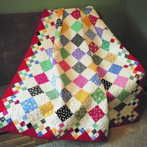 May include: A colorful handmade quilt with a diamond pattern. The quilt features squares in various colors, including red, pink, green, and blue, set against a white background. The edges are trimmed with a red border. The quilt is draped over a couch.