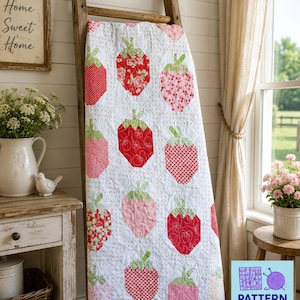 Op de afbeelding: Een witte quilt met een herhalend patroon van aardbeien in verschillende tinten rood en roze met groene steeltjes. De stof heeft een doorgestikte textuur met wervelende patronen. Een logo toont Pattern Warehouse Quilting Crochet Longarm.
