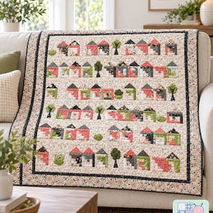 Op de afbeelding: Een rechthoekige quilt met een herhalend patroon van kleine huisjes en bomen in tinten rood, groen, zwart en wit. De quilt heeft een bloemenrand en een middengedeelte met rijen huisblokken. Een logo in de hoek toont Pattern Warehouse.