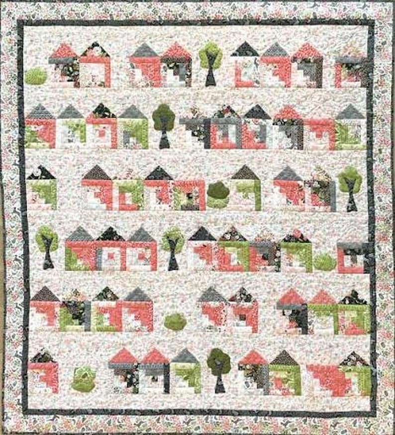 Op de afbeelding: Een rechthoekige quilt met een herhalend patroon van kleine huisjes en bomen in tinten rood, groen, zwart en wit. Het ontwerp is gerangschikt in horizontale rijen tegen een lichte bloemenachtergrond, afgewerkt met een donkere bloemenrand.