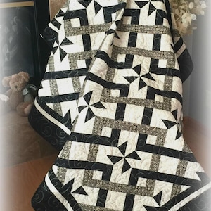 Op de afbeelding: Een zwart-wit quilt met een geometrisch sterrenpatroon. De quilt heeft afwisselende blokken van zwarte en witte stof, met een rand van zwarte stof met een wervelend ontwerp. De quilt is over een stoel gevouwen.