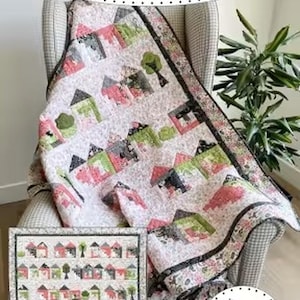 Op de afbeelding: Een quilt getiteld Tiny House Craze met een afgewerkte maat van 165,1 cm bij 177,8 cm. De quilt heeft een herhalend patroon van kleine huisjes in roze, groene en zwarte tinten op een lichte achtergrond, met een donkere rand en Jillily Studio merk.