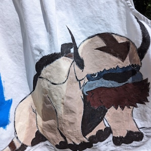 May include: White denim vest featuring a hand-painted image of Appa, a bison-like creature from Avatar: The Last Airbender. The artwork includes shades of brown, tan, black, and blue. The vest has a classic design with a pleated back.